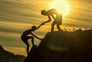 Color photo of a climber helping another climber up a mountridge.