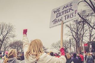 At the center of a group of protesters, a woman raises a fist and a sign that reads “WE ARE BETTER THAN THIS!”