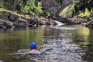 A swimmer in a blue hat swims along a sunny river as another swimmer sits under a bridge on a stone bank