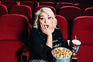 A blond woman in a red upholstered theater seat holds her hand to her mouth in astonishment while holding a bucket of popcorn and a soft drink.