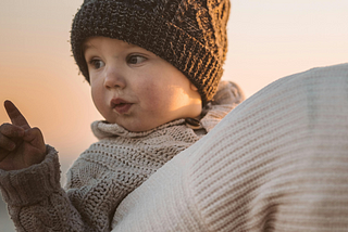 Toddler is bundled in crocheted hat and knit sweater and looking over mom’s shoulder who is carrying him and walking away from the camera. They are both bundled for the snow.