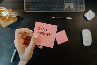 Manicured hand holding a post it reading “Don’t forget” above a computer keyboard and mouse