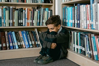 A schoolboy hugs his knees sitting in the corner of a library.