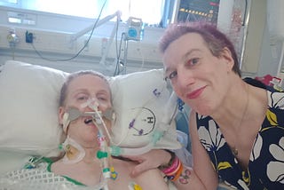 Abbie posing for the camera at Fae’s bedside, hand on Fae’s shoulder. Fae is breathing through medical equipment.
