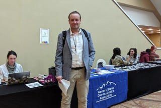 A man in a suit standing and facing the camera in front of a booth outside a convention.