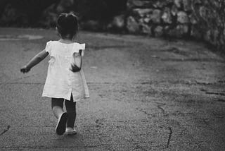 Little girl in a white dress and white shoes walking on a paved road with her back turned to the camera. Her hair is in a ponytail hair-do and some hedges are in the background.