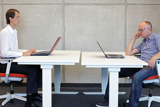 two men sitting across from each other at an office with laptops and one is staring creepily at the other one