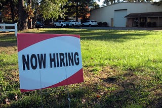 A lawn sign that reads “Now Hiring”.