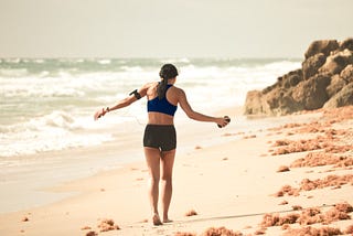 Young Woman walking along a rocky beach, wearing sport shorts and a sports bra, with a phone and headphones. Dark long hair in a low ponytail. Daytime.