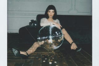 Woman sits on the floor behind a giant disco ball, looking solemn.