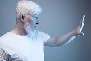 A serious mature bearded male with gray hair doing protective gesture while standing in a white light