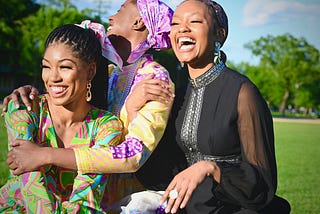 A trio of young black women laugh and embrace while sitting outside.