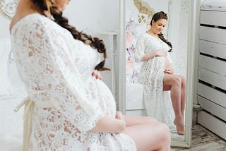 Pregnant woman wearing white sitting on edge of bed reflected in mirror
