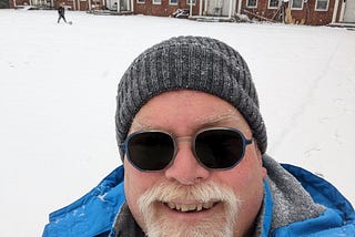 Selfie of the author, John Carney, wearing a stocking cap and heavy winter coat and standing in front of his apartment building. The lawn is covered with snow. Carney’s light-sensitive eyeglass lenses have fully darkened.