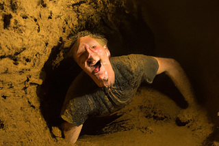 DALL-E generated image shows a person “having fun” in a cave — squeezeing through a small gap surrounded by mud.