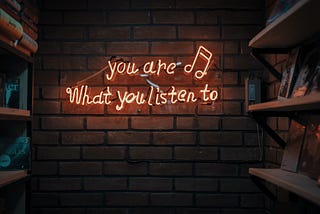 photo of text ‘you are what you listen to’ in neon lights aside music notes hanging in brick wall. Photo on Dr. James Goydos 2021 article on physician satisfaction podcasts