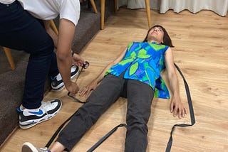 A photo of a person in a blue shirt lying on the floor. Another person in a white top lays duct tape around her body, in the shape of her body.