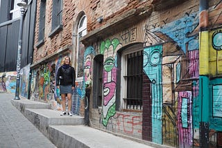 A woman stands in front of a colorful mural of street art in Valparaíso, Chile. She is smiling and has blonde, shoulder-length hair and is wearing a black jacket, a dress, and black sneakers.