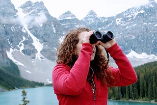 Woman with binoculars looking at something in distance with mountains and a lake behind her.