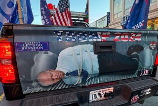 The tailgate of a pick-up truck. Text across the top reads Trump 2024, with a ‘Let’s Go Brandon’ bumper sticker to the left. Below is an image of President Biden hogtied in the bed of the truck.