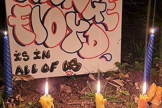 A small memorial poster surrounded by candles reads “George Floyd is in all of us”