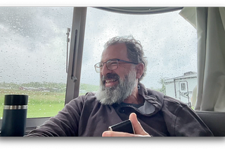 The author smiles while sitting inside an Airstream on a rainy day, holding a phone during a recording.