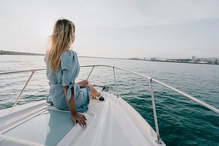 Woman in a blue dress sitting on the bow of a boat at sea during daytime.