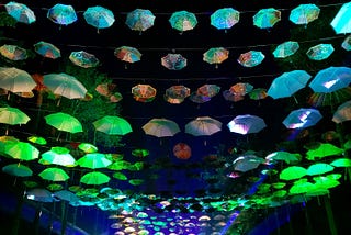 Groups of people gathered together to sing and dance and do human things, with umbrellas lining the “ceiling” of the clearing in the trees, and lights coming from the far end.