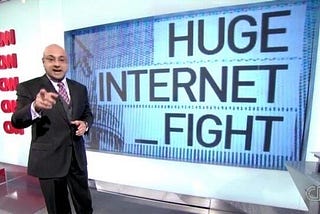 A screencap of a news desk. The reporter is pointing at the camera. To his right, a big screen with the words “HUGE INTERNET FIGHT” is shown.