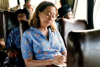 Mulher branca, cabelos castanhos claros, está sentada no ônibus de viagem e está escrevendo em um papel. Ela veste uma camisa azul com estampa de flores na cor rosa.