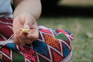 close-up on a person sitting on the ground wearing a brightly colored skirt pulled over the knee, with hand resting on the leg. In the hand is a small, remaining bite of a crispy, protein bar.