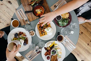 A round table with multiple plates full of breakfast food. There are two people sat at the table. One is reaching over, the other is lifting a cup of coffee.
