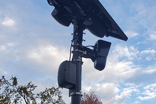 A black pole topped with a solar panel, and two oblong devices attached with wires against a cloudy sky.