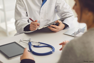 Profissional da saúde, usando jaleco branco, conversa com uma paciente em um consultório. O médico segura uma prancheta com papéis e caneta, enquanto a paciente está sentada à frente. Sobre a mesa estão um estetoscópio azul, um tablet e um notebook