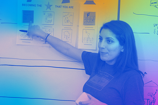 Tania Anaissie stands near a white board and points to a handout taped to its surface. The title of the handout says, “Becoming the Star That You Are.” This image is colorized with shades of light blue, gold, green and pink.