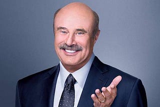 A professional profile photograph of Dr. Phil in a blue suit and tie, and crisp white shirt. He is posing with a smile and his hand is held out with his palm opened upward as a friendly looking gesture.