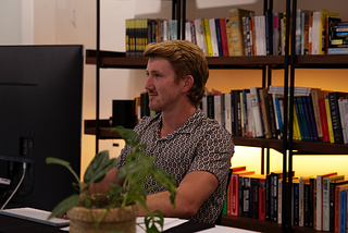 man sitting in front of a computer writing PyTorch code, there is a bookcase behind him and a plant in front of him