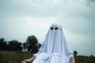 Woman sitting on grass, wearing snickers and covered with a white sheet from head to toes, wearing sunglasses over it