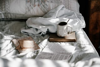 A cup of a beverage sits on a book on top of a mattress with white bedding and scattered books and journals, opened and closed lay around it.
