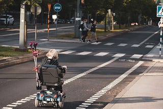 A person on a mobility scooter is riding down the empty street on a dedicated lane towards a crossroads. Two people are about to cross the street on a pedestrian crossing