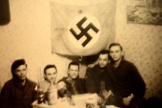 295th men (Rogers, Deragio, Franklin, Sage, and Jack Hope) sitting at a kitchen table with Jack’s captured Nazi flag.