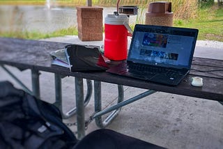 a park bench and table with a laptop, giant red jug with a straw, a seat cushion, backpack and binders with water and a fountain in the background, surrounded by grass and trees