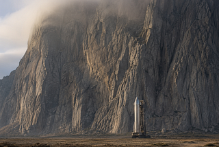 A gleaming modern rocket stands dwarfed at the base of an immense granite cliff that fills the frame, symbolizing human ambition and capital confronting the immovable laws of physics.