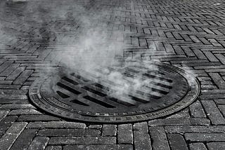 A sewer grate in black and white. Steam exits the grate.