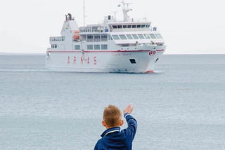 Young boy dressed in blue waving goodbye to a cruise ship.
