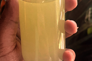 A woman’s hand holding a small glass spray bottle that has yellow liquid in it that looks like lemonade. A plant is in the background.