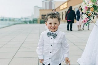 A little boy wearing a bow tie stands on a sidewalk, smiling and looking towards the camera.