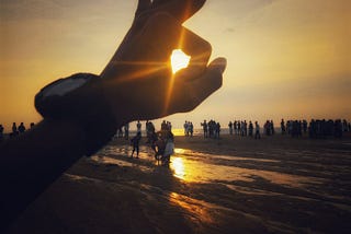 A setting sun captured in the middle of a hand making an okay sign with a beach background.