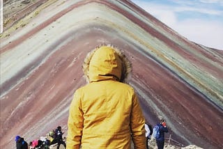 A person in a mustard yellow fur-lined hood stands facing Peru’s Rainbow Mountain, with its vibrant natural stripes of red, green, and gold layered across the peak under a partly cloudy sky. Other hikers rest and take photos in the background.