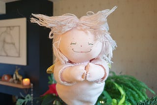 Photo of a pink Christmas tree fairy. She has the most beautiful smile and her hands are clasped together in front of her. Her hair is made of pink cotton threads and it is sticking out at funny angles. The blurred background shows green foliage, a poinsettia, a candle and other Christmas decorations.